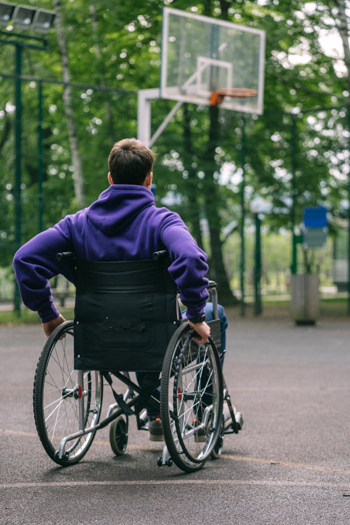 A man in a hoodie plays basketball on an outdoor court from a wheelchair, promoting inclusivity.